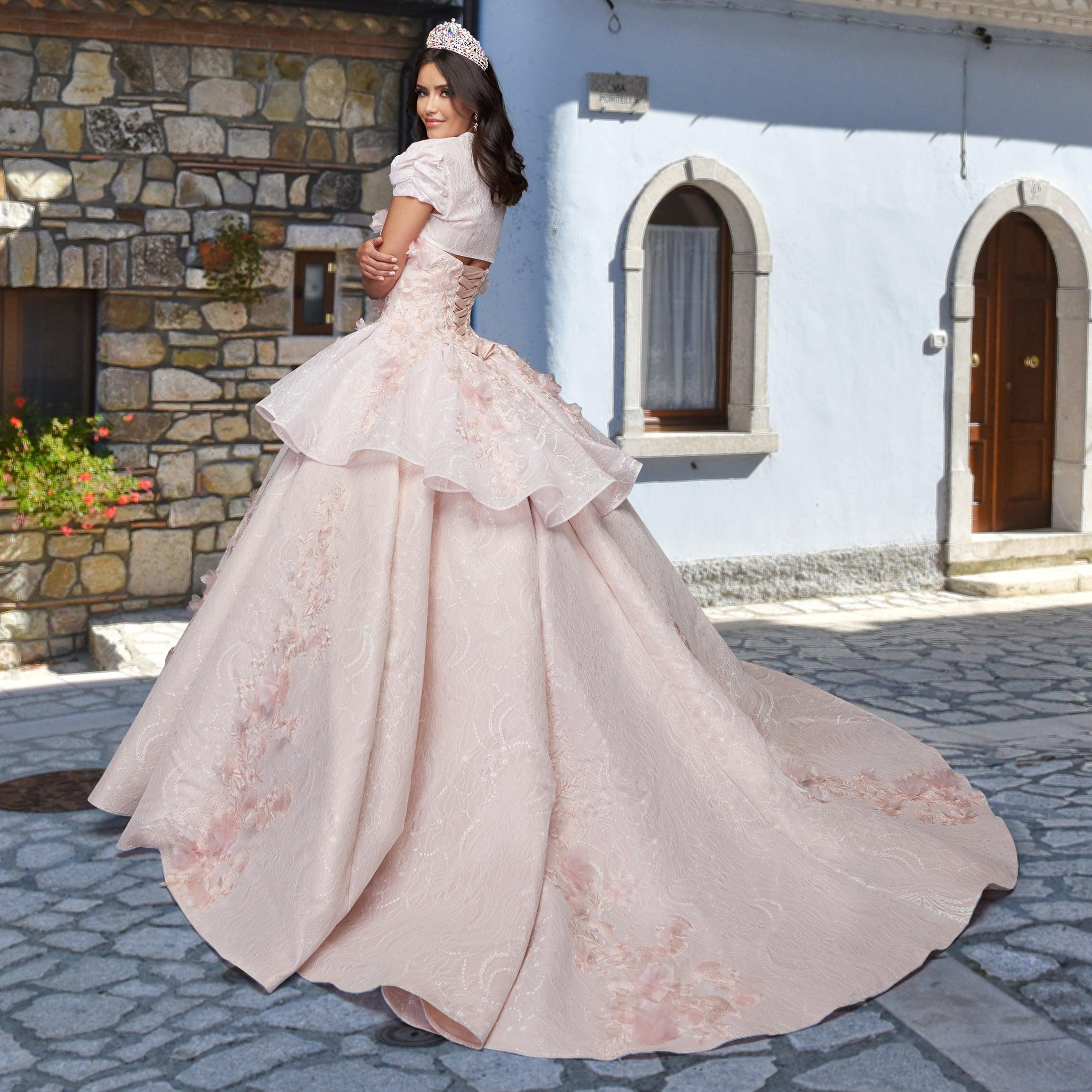 A woman wearing the Quinceañera Sample Dress QF115 and a tiara stands on a cobblestone street before a light blue building with arched windows and doorways.